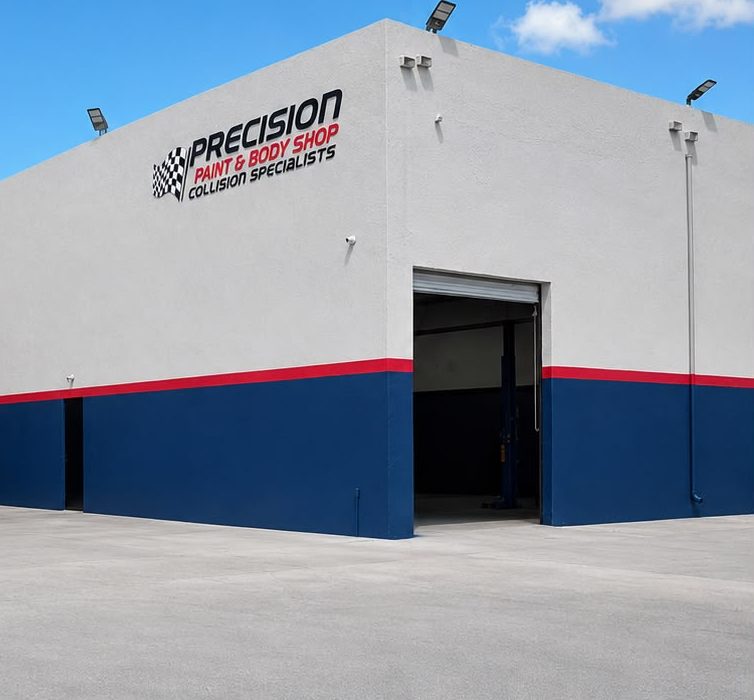 An open repair bay at the Opa-locka shop, with a vehicle lift visible inside the doorway and the Precision Paint & Body Shop name on the wall above